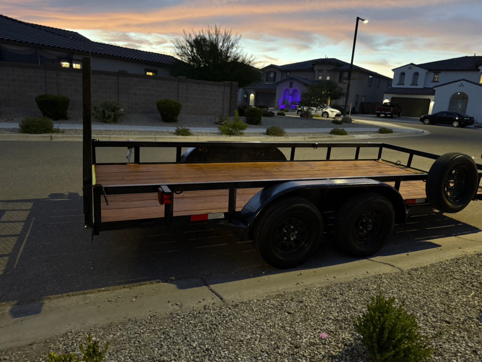 16 foot black tandem-axle utility trailer with wooden deck and rear ramp door, parked on a residential street at dusk.